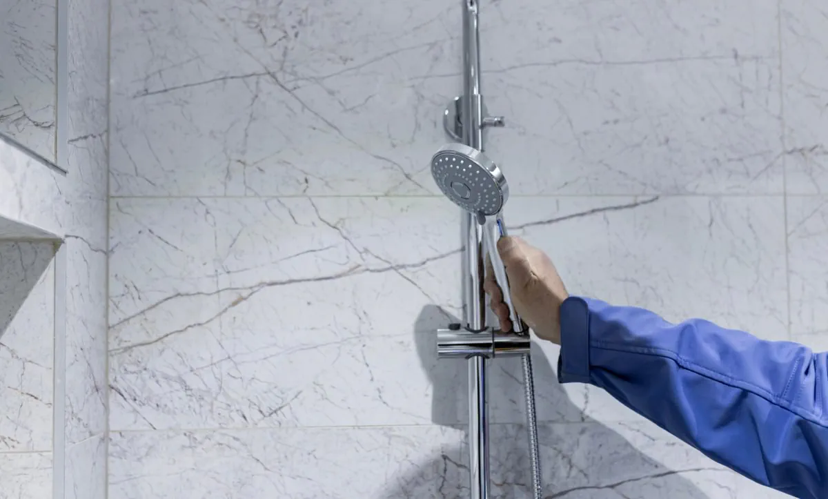Man adjusting a handheld showerhead in a marble-tiled bathroom.