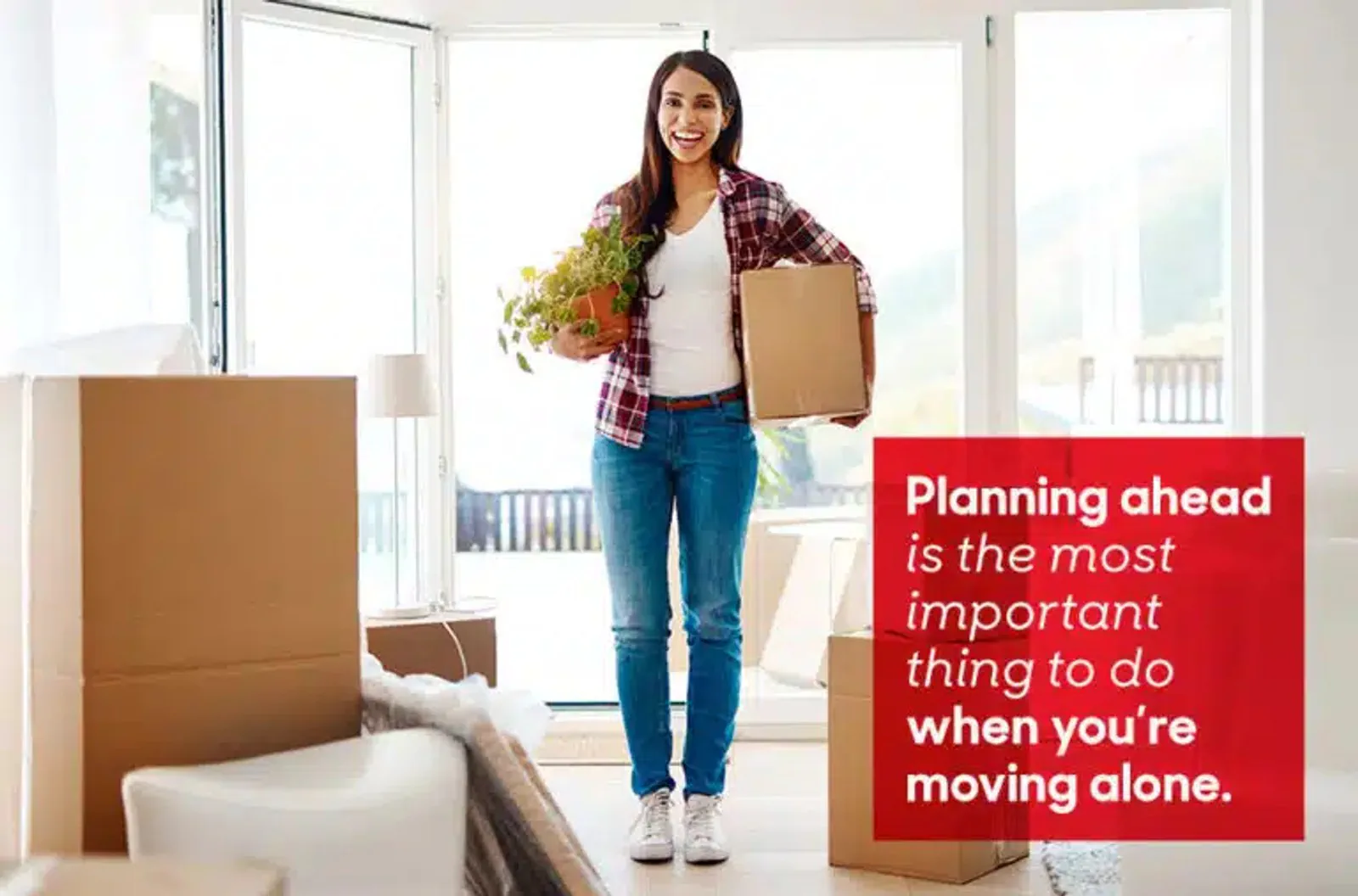 A woman carries a moving box and a houseplant through a bright room.
