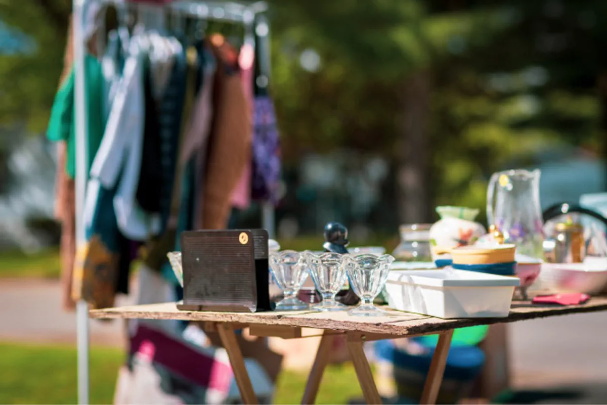 A moving sale, featuring a makeshift table covered in miscellaneous items with price stickers on them and a rack of clothing.