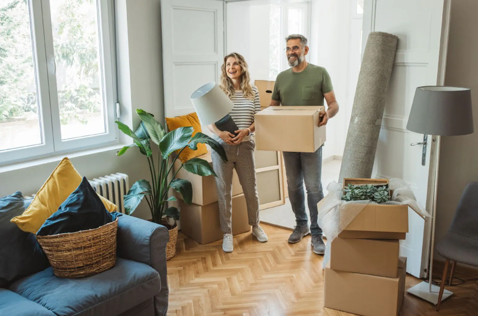 A couple carries boxes and a lamp while preparing a home for a move.